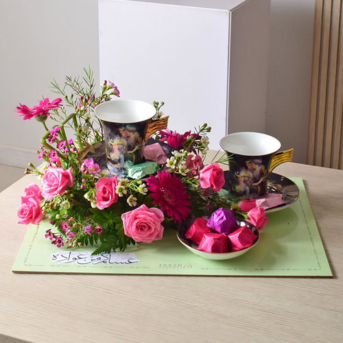 Me & You 1 - 2
  ceramic coffee cups set with 1 ceramic plate of chocolates with a decoration
  of fresh gerbera, roses and wax flowers on a board tray.