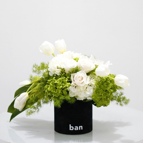 Eirwen - A bouquet of white with touch of greens. White tulips cymbidium, roses hydrangeas in black velvet cylinder box, H 30 x W 45 cm