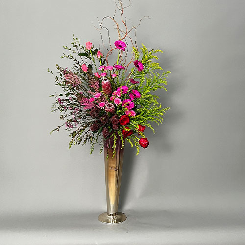 Passion - Pink and gold long vase with fuchsia gerbera, pink rose, wax, pink baby rose, red protea, skimmia and red roses  
 Height 140  Width 65