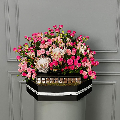 The Garden - A wooden basket with light and dark pink baby roses and protea with Belgian chocolates. 
 Height 55  width 65