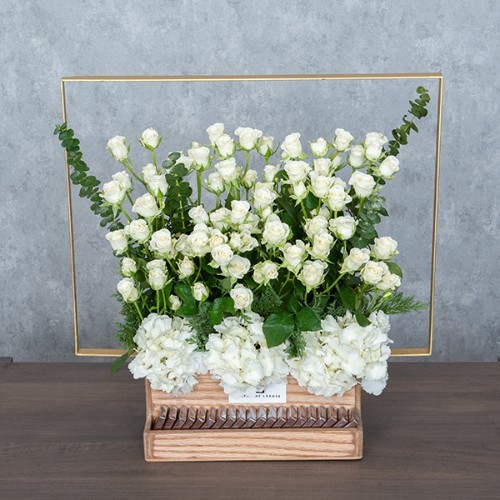 Fluffy White - A wooden basket with golden metal handle contains white baby roses, hydrangea and eucalyptus with Belgian chocolates.  Height: 52cm – Width: 51cm