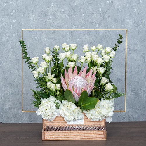 Fluffy White II - A wooden basket with golden metal handle contains king protea, white baby roses, hydrangea and eucalyptus with Belgian chocolates. Height: 52cm – Width: 51cm