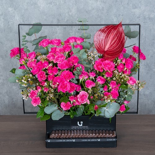 Sybya II - A black wooden basket with metal handle contains baby roses, anthurium, white wax, and eucalyptus with Belgian chocolates.  Height: 55cm – Width: 57cm