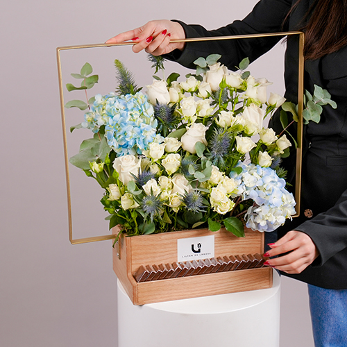 Sophia - This wooden basket contains blue hydrangea, baby roses and eucalyptus with Belgian chocolates. Height: 51 cm - Width: 51 cm