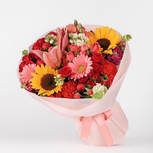 La Rosée du Soleil - This bouquet appears to include a romantic and vibrant mix of the following blooms: 1. Sunflower • Symbol: Positivity, strength, and admiration • Adds a bold, golden centerpiece to the bouquet. 2. Asiatic Lilies (Pink) • Symbol: Prosperity and abundance • Brings elegance and a touch of femininity. 3. Gerbera Daisies (Pink/Purple) • Symbol: Cheerfulness and innocence • Adds playful color and round texture. 4. Roses (Deep Red) • Symbol: Passion, love, and elegance • Deepen the luxury and romantic theme. 5. Carnations (Pale Pink/Peach) • Symbol: Gratitude and admiration • Provides fullness and softness. 6. Stock Flowers (Light Lavender/White)