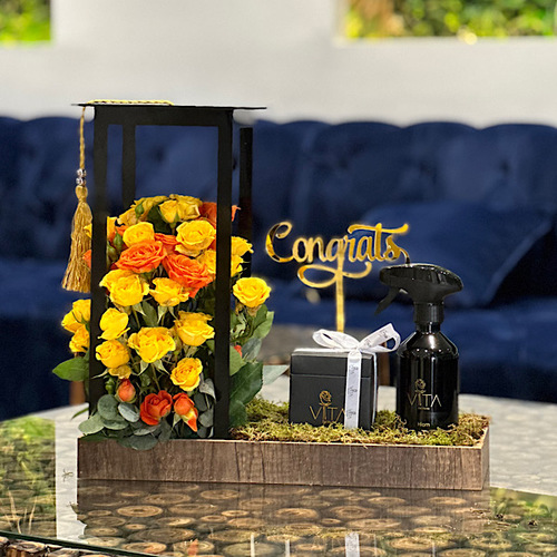 Copra - Wooden tray contains yellow & orange baby roses, green leaves, with graduation cap, graduation gold stick and spray perfume and box of chocolates. Height: 55 Width: 35