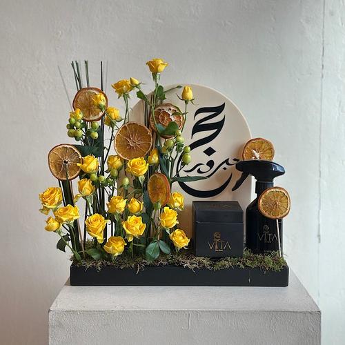 Ero - A tray elegantly filled with steel grass, hypericum green, yellow baby roses and dried fruits with a box of chocolates and a home fragrance spray. Height: 40 Width: 20