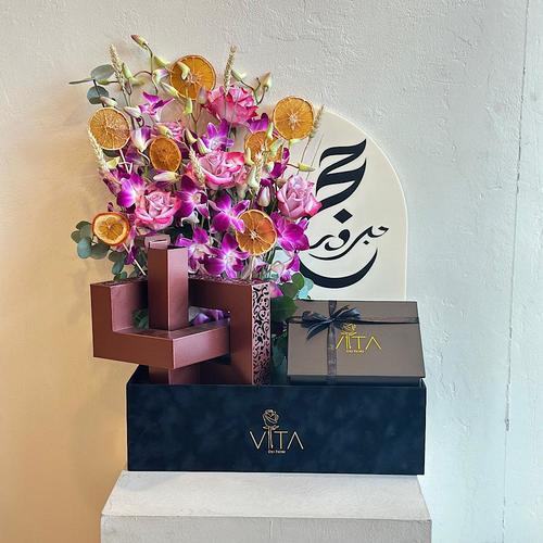 Kos - A black box contains purple baby orchid, deep purple rose,dried fruit, mubkhar with a chocolates and acrylic hajj topper. Height: 70 Width: 40