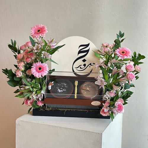 Marousi - A tray elegantly filled with baby roses, gerbera and green leaves with with a middle acrylic box containing mubkhar and charcoal handle with an empty box and acrylic hajj topper. Height: 40 Width: 20