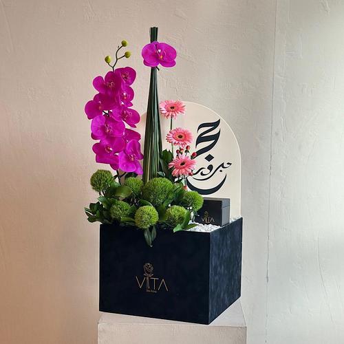 Volos - A box filled with purple orchid cut flowers, steel grass, gerbera, hypericum and green leaves whit a chocolates and acrylic hajj topper. Height: 70 Width: 40