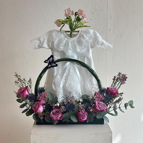 Arges - A tray that contains purple roses with eucalyptus leaves, steel grass, eryngium, and wax flowers. Comes with a white dress for a newborn baby. Height: 60 Width: 20