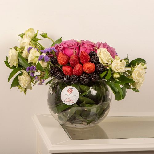 Medium Round Vase 503 - A round transparent vase that contains 5 purple roses, 5 white baby roses, 2 purple statics, 5 green ruscus, fruits: 6 strawberries, 10 blackberries
height 25 width 40.