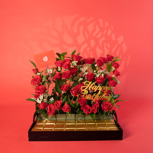 Baby Red baby Roses and wax Flowers, and Ruscus With Wafer Chocolate in acrylic tray 1702 - An elegant arrangement of premium red roses accented with delicate white blooms and fresh greenery, artfully styled on a sleek base. Presented with a row of gold-wrapped chocolates for a luxurious touch. A refined gift for any special occasion that blends beauty and indulgence. dimensions  Length 35 cm Width 25 cm Hight 27 cm